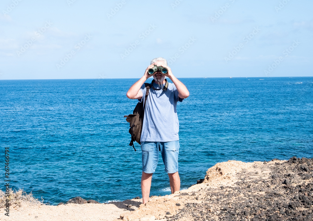 Elderly man white hair standing on the cliff with a beautiful seascape. Adult 70 years old. Blue background. One people looking with a binoculars