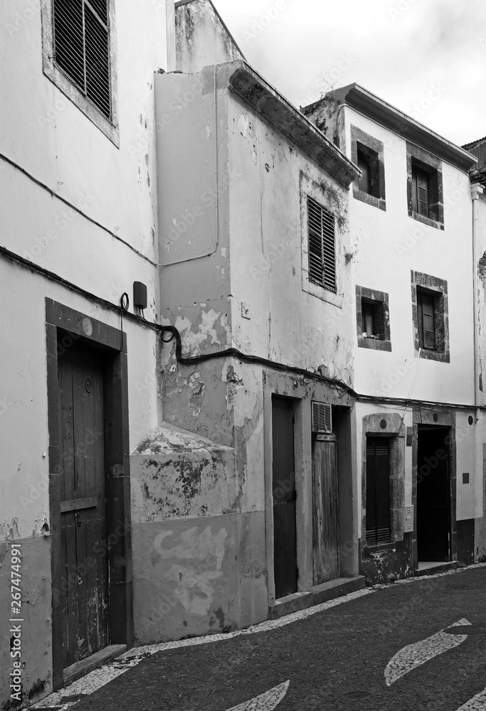 monochrome image of a typical quiet empty street in funchal madeira with old traditional houses painted in faded peeling paint and a cobbled road