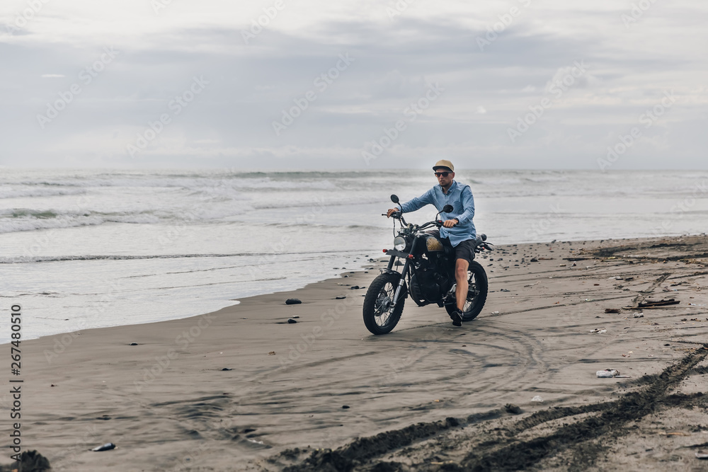 Obraz premium Man in cap riding motorcycle on beach. Moto cross dirtbiker on beach sunset on Bali. Young hipster male enjoying freedom and active lifestyle, having fun on a bikers tour.