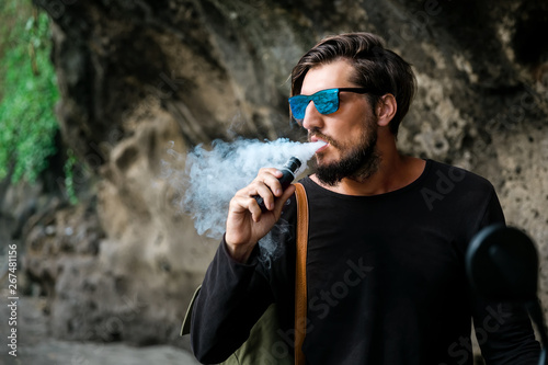 Man with beard and sunglasses seat on motorcycle smoking electronic sigarette outdoor on sunset beach. Young hipster male vaping on sea front.