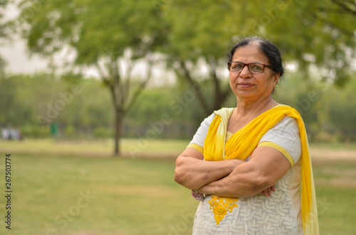 Smart senior north Indian woman standing, posing for the camera with hands crossed in a park wearing yellow salwar kameez in summers in Delhi, India