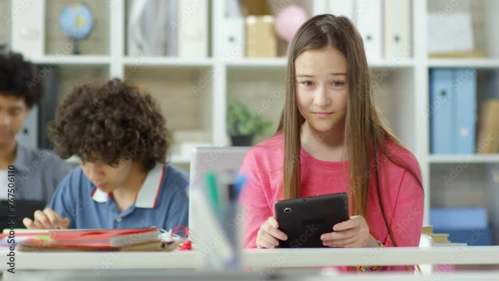 Tracking right of several students sitting at school desks with tablets in their hands. One girl with strait long hair looking at camera and smiling