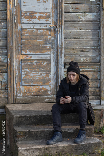 European teenager with dreadlocks in a black hat and clothes sitting on the steps of an abandoned house. Close-up