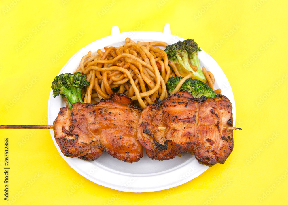 White paper plate with teriyaki chicken on skewer with chow mein noodles on yellow table cloth. Popular street fair food.