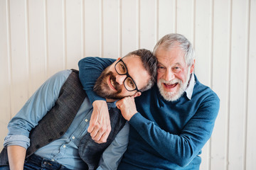 A portrait of adult hipster son and senior father sitting on floor indoors at home.
