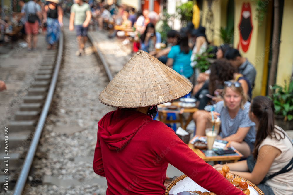 Vietnamese woman vendor walking along railroad with people drink coffee or walking on railways waiting for train to arrive on railway road in Hanoi, Vietnam. Vietnamese woman vendor walking along railroad with people drink coffee or walking on railways waiting for train to arrive on railway road in Hanoi, Vietnam.