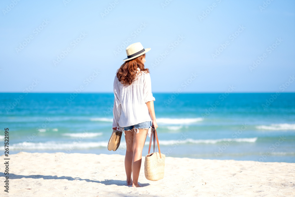 A woman holding bag and shoes while strolling on the beach with the sea and blue sky background