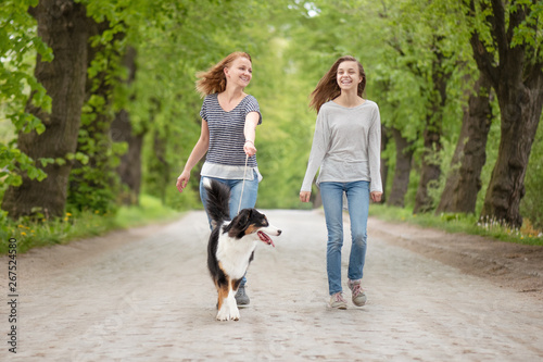 Happy family - mom and daughter, walking with dog. Woman and girl with Australian shepherd dog. Mother and child go on road in city park. They are talking and enjoying beautiful day.