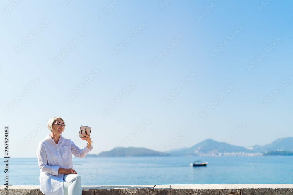 Smiling mature woman having a video chat through tablet on background the sea and blue sky