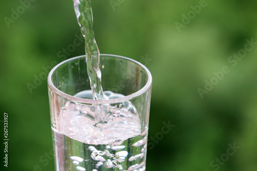 Pouring clean water into drinking glass on green nature background. Concept of health and freshness, thirst, water purification and ecology