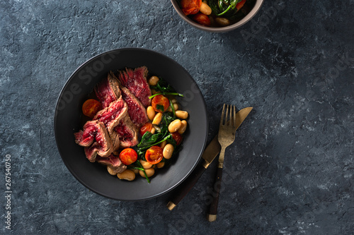 Dinner table. Beef steak with white beans, spinach and tomatoes on dark stone background top view with free copy space