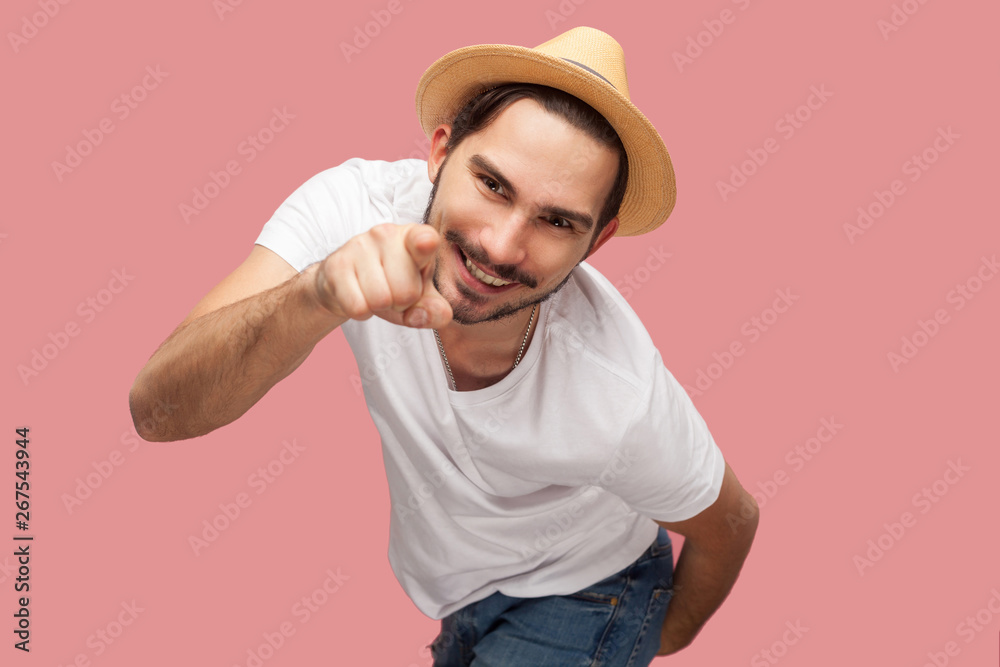 Portrait of funny bearded young man in white shirt with hat standing, pointing and looking at camera with toothy smile. indoor studio shot, isolated on pink background copyspace. Portrait of funny bearded young man in white shirt with hat standing, pointing and looking at camera with toothy smile. indoor studio shot, isolated on pink background copyspace.