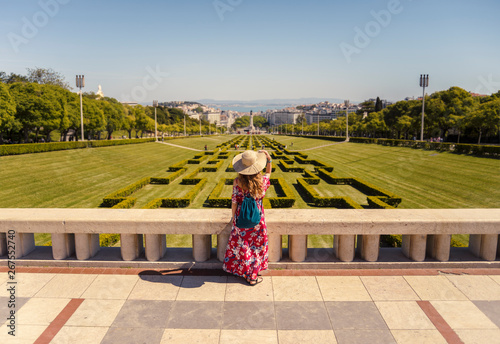Young blonde woman wearing red generic sundress and a hat looking at cityscape view of Lisbon, capital city of Portugal