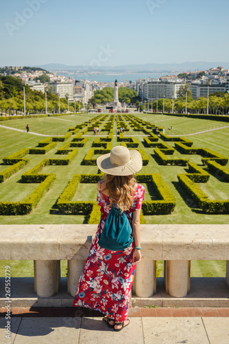 Young blonde woman wearing red generic sundress and a hat looking at cityscape view of Lisbon, capital city of Portugal