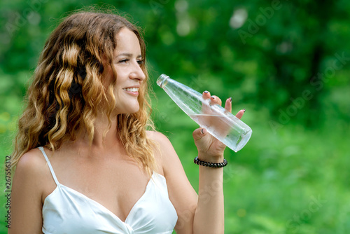 Beautiful young woman drinking fresh clear water from a glass bottle on nature background