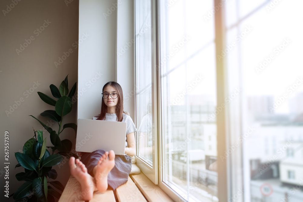 Cute teenage girl sitting on window sill at home and using laptop. Education, children and school concept, distance learning, self education Cute teenage girl sitting on window sill at home and using laptop. Education, children and school concept, distance learning, self education