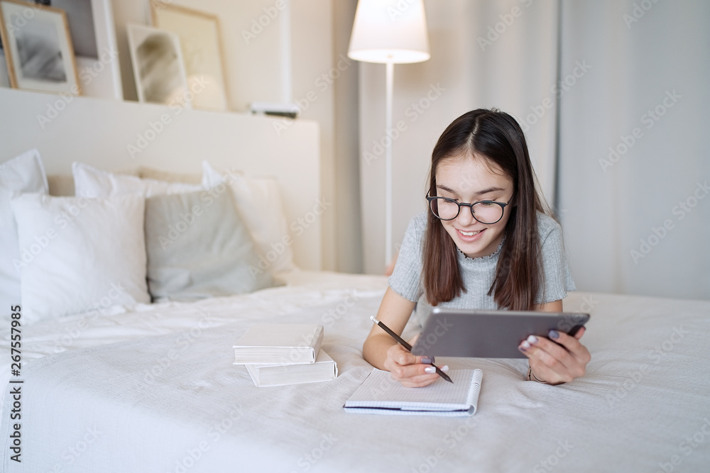 Cute teenager girl doing homework lying on bed at home. Young pretty girl wearing glasses writing down in notebook, studying online with tablet, distance learning, self education Cute teenager girl doing homework lying on bed at home. Young pretty girl wearing glasses writing down in notebook, studying online with tablet, distance learning, self education