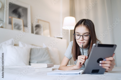 Cute teenager girl doing homework lying on bed at home. Young pretty girl wearing glasses writing down in notebook  studying online with tablet  distance learning  self education     