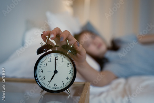 Close up sleepy teenager girl turning alarm off in the morning while lying in bed. Early wake up, not getting enough sleep, getting work concept photo