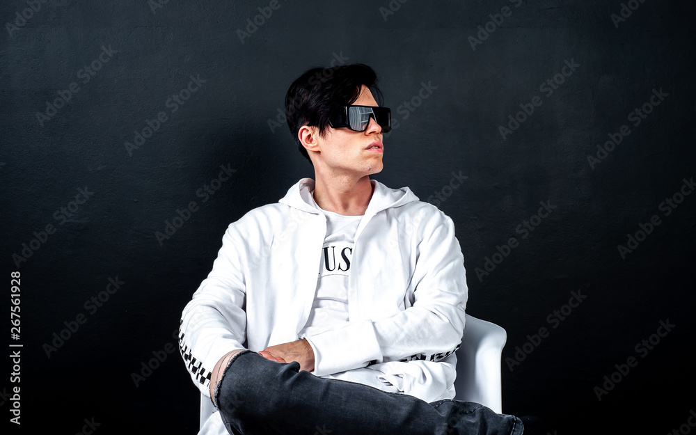 a guy in black glasses is sitting on a chair against a dark background of concrete in a studio; in the studio is a young guy in black glasses