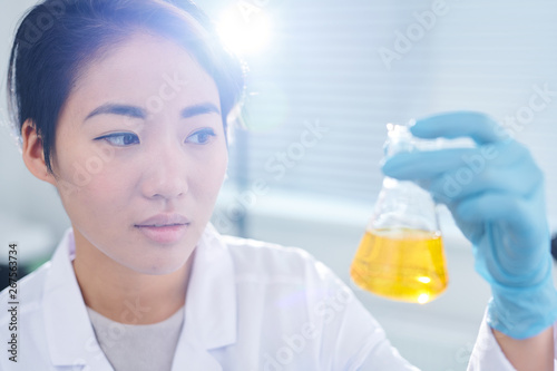 Serious puzzled young Asian laboratory worker in rubber gloves standing in sunlight and examining chemical liquid in flask