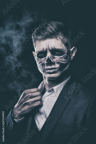Stylish and beautiful, emotional young man with skeleton makeup in a strict suit against the background of smoke and dark background to Halloween