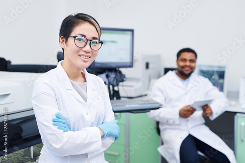 Smiling attractive smart young Asian female medical scientist in white coat and disposable gloves standing in laboratory and crossing arms on chest