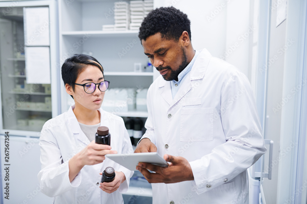Serious thoughtful multi-ethnic pharmacists in lab coats standing in drugstore and discussing new medications while using tablet to search for information in database Serious thoughtful multi-ethnic pharmacists in lab coats standing in drugstore and discussing new medications while using tablet to search for information in database