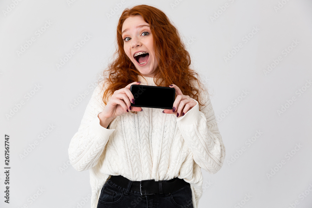 Beautiful young girl redhead posing isolated over white wall background showing display of mobile phone.
