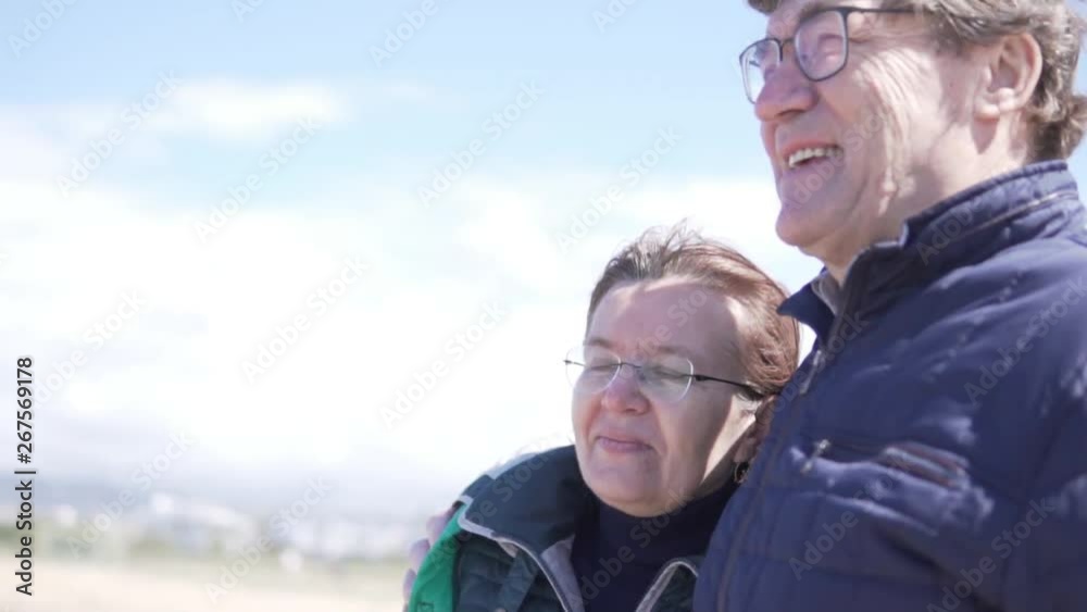 A happy elderly couple is looking at the sea. A man and a woman with glasses snuggle up to each other and smile, looking at the ocean. Walking retirees in Cyprus in the winter