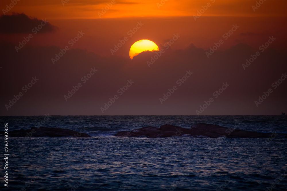view of the sun partly hidden behind clouds and the sea with rocks.