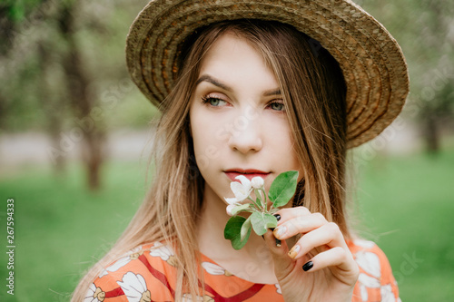 Young beautiful blonde woman in blooming garden. Spring trees in bloom. Orange dress and straw hat.