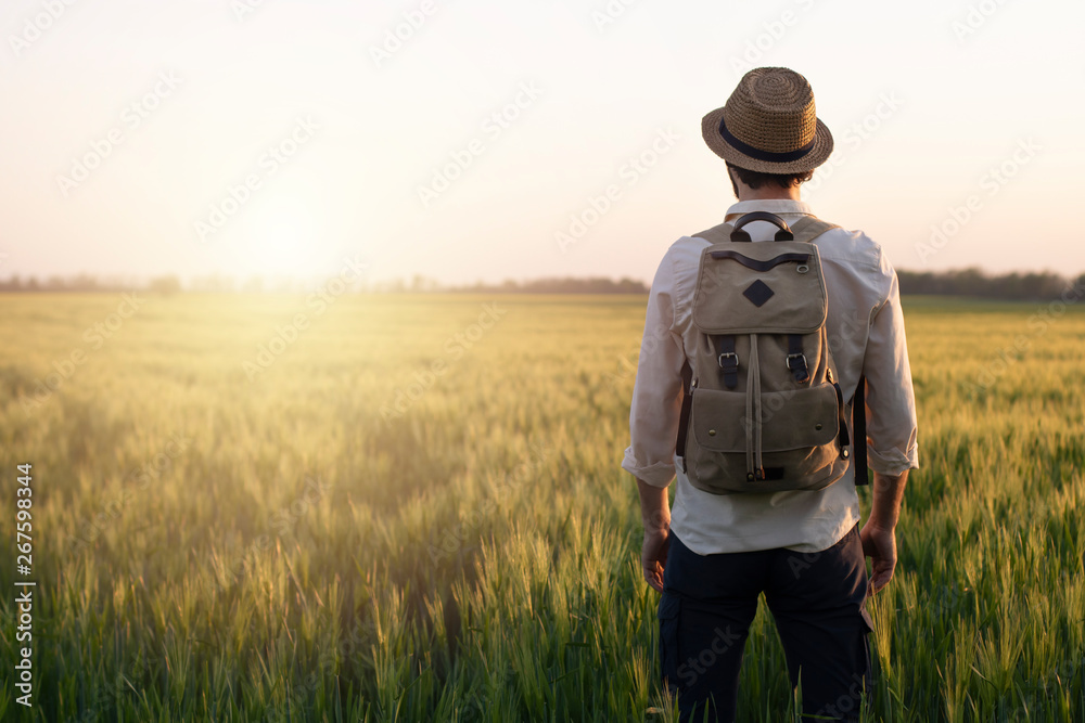 Traveler with a backpack in a field of wheat at sunset. Man outdoor in nature at the backdrop of the open horizon. The concept of freedom and discovery. Environment and lifestyle