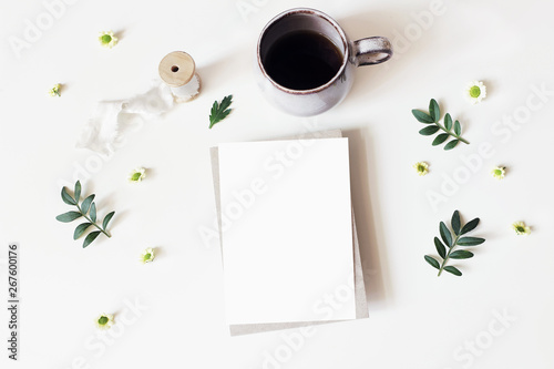 Floral lifestyle, stationery scene. Santini Chrysanthemum flowers, lentisk leaves and cup of coffee on white table. Blank greeting card mockup. Rustic design. Flat lay, top view.