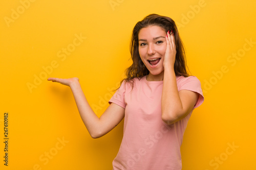 Young european woman isolated over yellow background holds copy space on a palm, keep hand over cheek. Amazed and delighted.