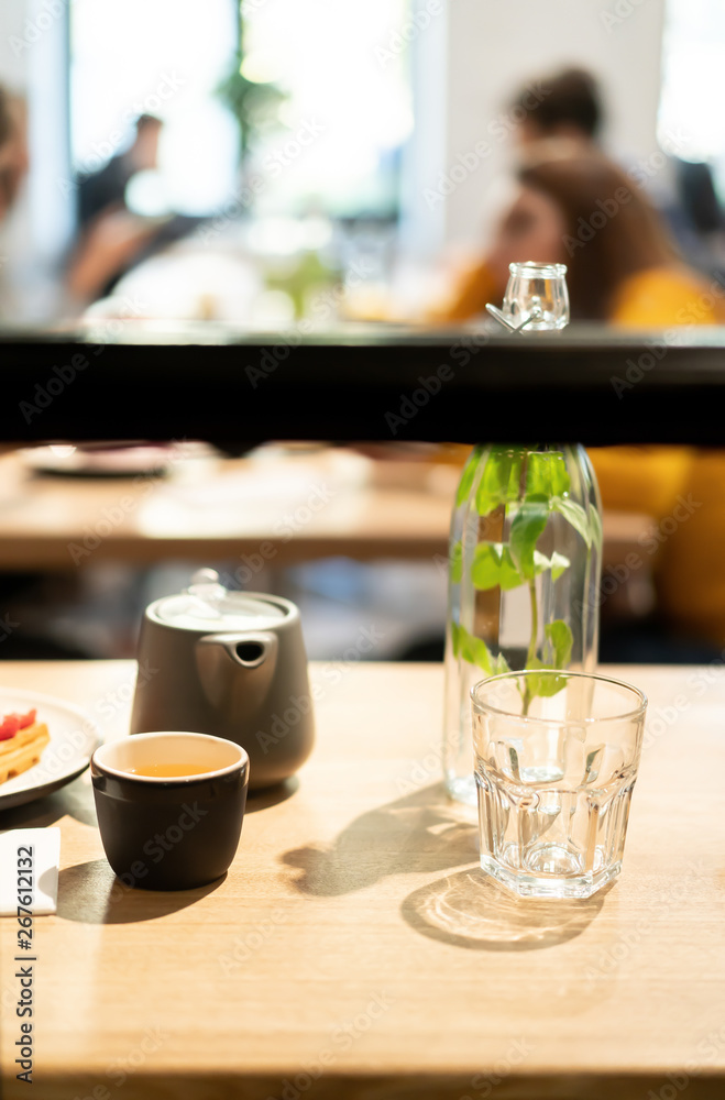 Gray teapot on a wooden table in a cafe. Black mug for tea. Dessert on a white plate and sliced lemon on a small saucer. Glass for water and a carafe. Gray teapot on a wooden table in a cafe. Black mug for tea. Dessert on a white plate and sliced lemon on a small saucer. Glass for water and a carafe.