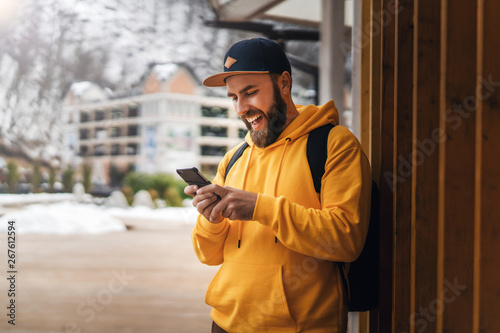 Front view. Bearded male tourist in yellow hoodie and cap with black backpack stands outdoors and using smartphone. Lifestyle.