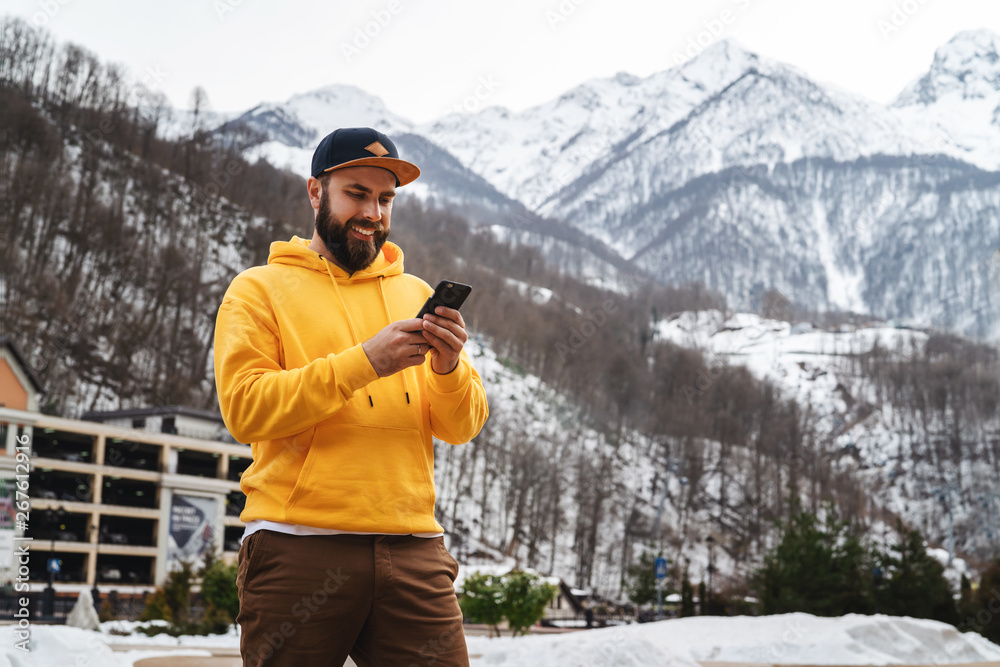 Front view. Man tourist in yellow hoodie, cap stands on background of high snowy mountains and using smartphone. Lifestyle.