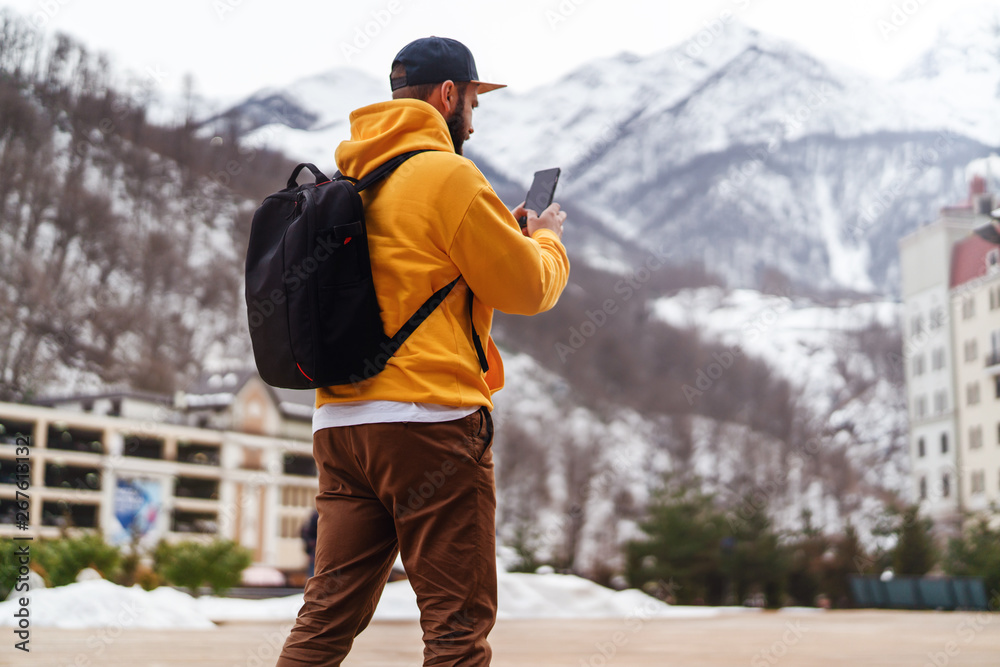 View from back. Man tourist in yellow hoodie, cap with backpack stands on background of high snowy mountains and using smartphone.