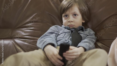 The little cute boy sitting on the leather sofa with legs apart changing channels on TV using remote. Cute child in shirt and tie resting at home. Kid as adult photo