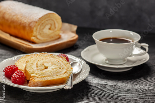 Ciasto - rolada biszkoptowa z kremem mascarpone na talerzyku, obok kawa w filiżance, wszystko postawione na czarnym drewnianym blacie