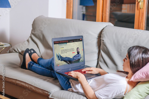Young woman lying comfortably on the sofa waching video streaming website on laptop. All screen graphics are made up. photo
