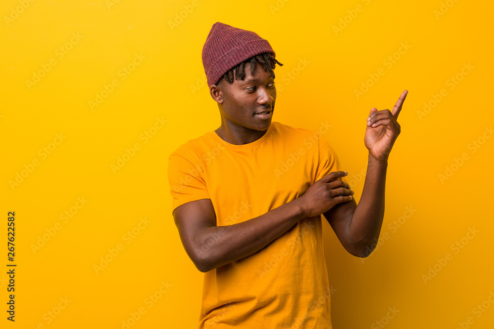 Young black man wearing rastas over yellow background smiling cheerfully pointing with forefinger away. Young black man wearing rastas over yellow background smiling cheerfully pointing with forefinger away.
