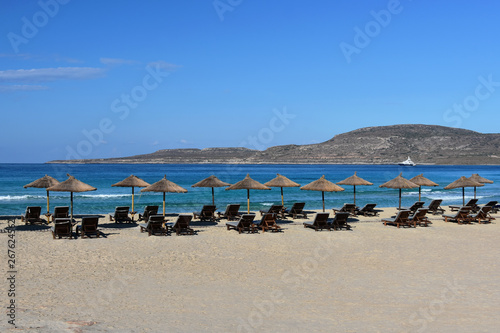 Wide sandy beach with old umbrellas and empty chaise longue  quiet turquoise sea  white yacht far away in the bay  serene blue sky  Simos Beach  Elafonisos Island  South Greece