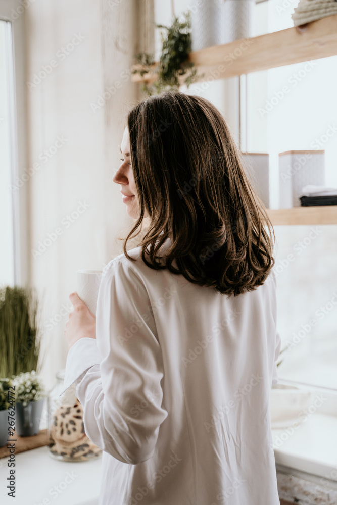 Successful and confident woman on a morning coffee break, resting and enjoying the drink. Young girl in white panties and long shirt with naked shoulder and chest stand on the kitchen of the house Successful and confident woman on a morning coffee break, resting and enjoying the drink. Young girl in white panties and long shirt with naked shoulder and chest stand on the kitchen of the house