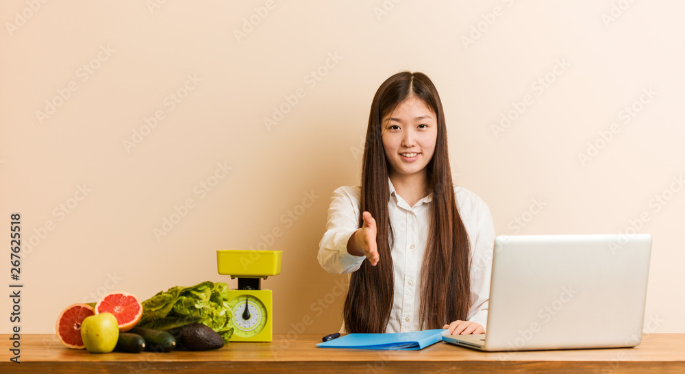Young nutritionist chinese woman working with her laptop stretching hand at camera in greeting gesture. Young nutritionist chinese woman working with her laptop stretching hand at camera in greeting gesture.