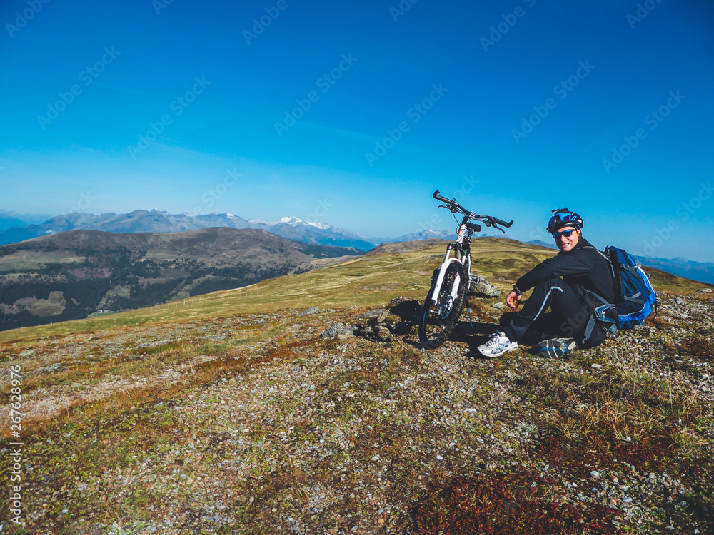 A young boy on a mountain bike tour in Millstatt, Austria. Boy wears a big backpack and a helmet. Mountain bike stands next to him. Beautiful mountain view in the back. Professional sport gear.