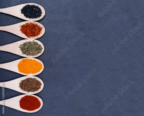 Collection of spices in wooden spoons (Saffron, Basil, Turmeric, Caraway seeds, Sweet Paprika, Black Lava Sea Salt) on dark background.