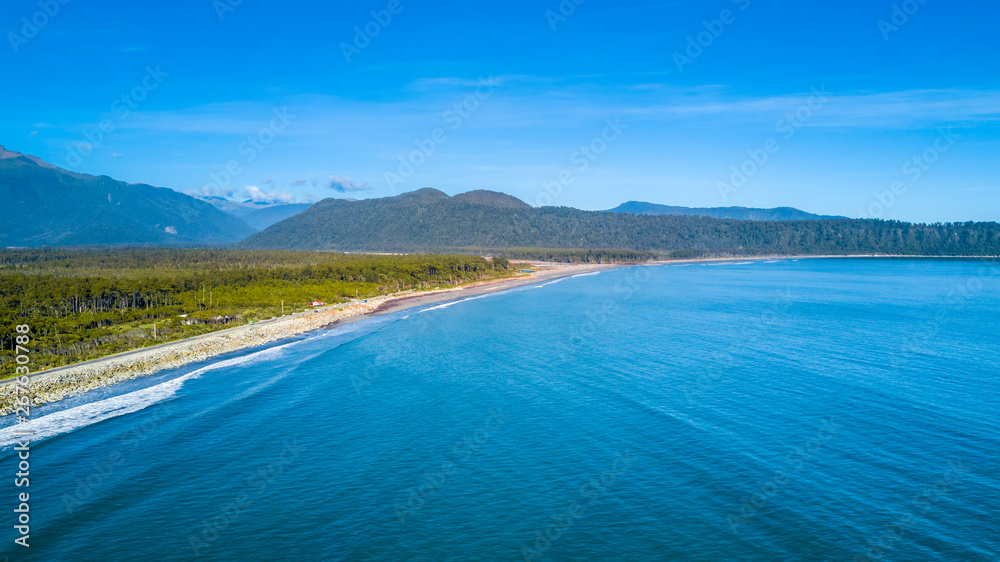 Road running along a sunny beach with a peninsula and mountains on the background. West Coast, South Island, New Zealand