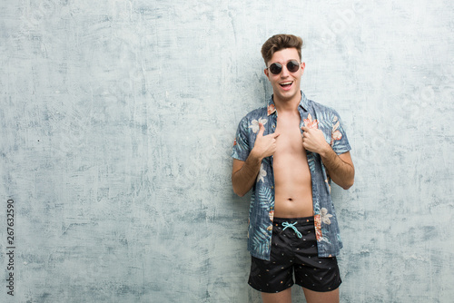 Young caucasian man wearing a swimsuit surprised pointing at himself, smiling broadly.